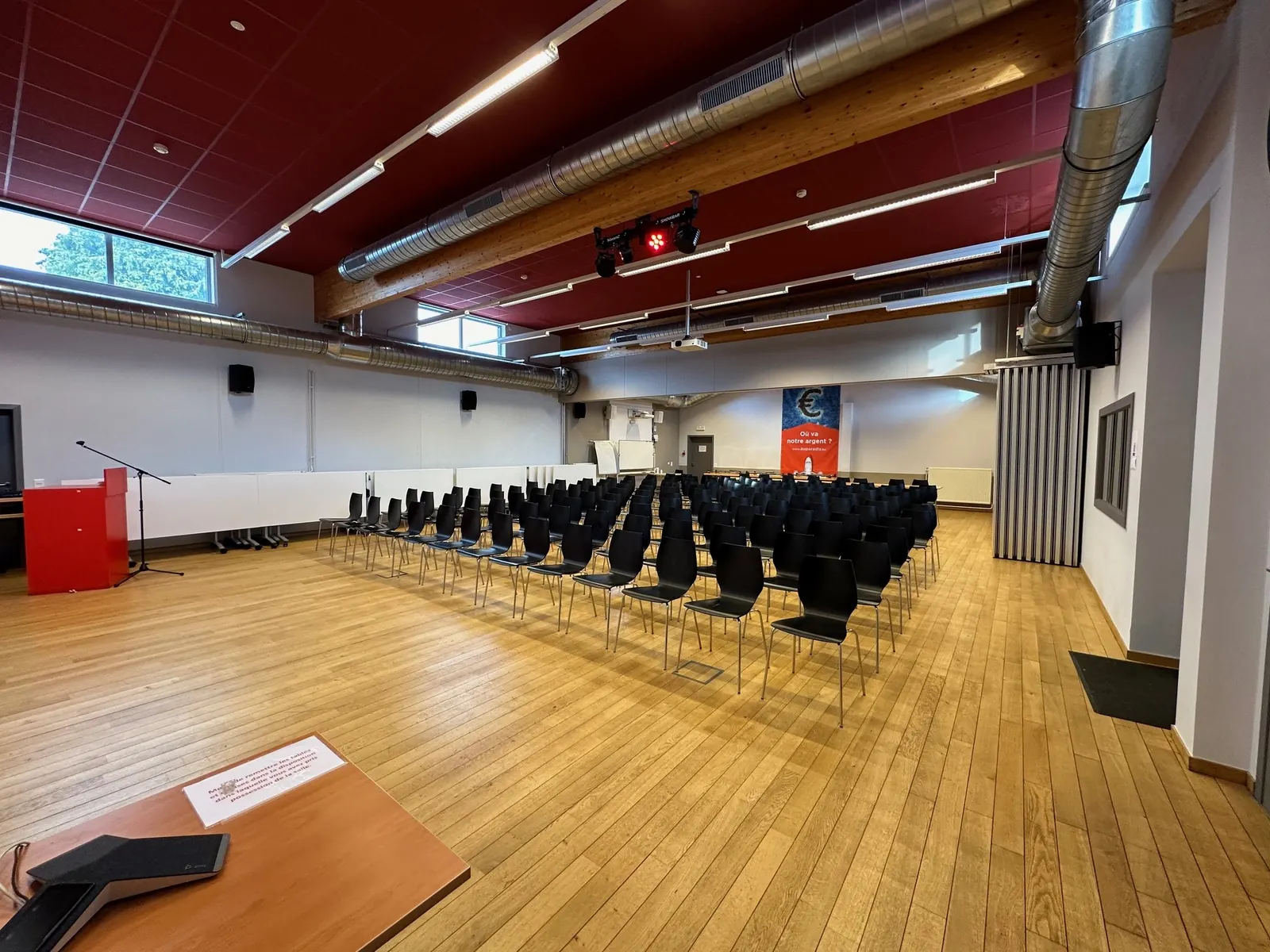 Vue large de la grande salle : rangées de chaises, pupitre, éclairage de scène suspendu au plafond rouge.