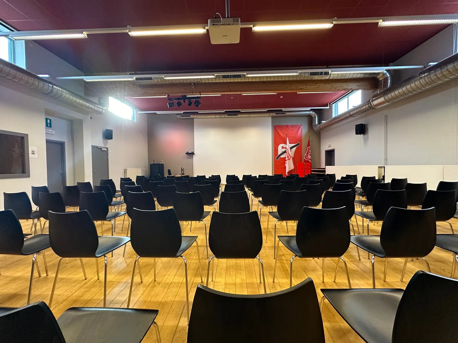 Grande salle vue depuis l'arrière, chaises alignées vers le pupitre et le drapeau.