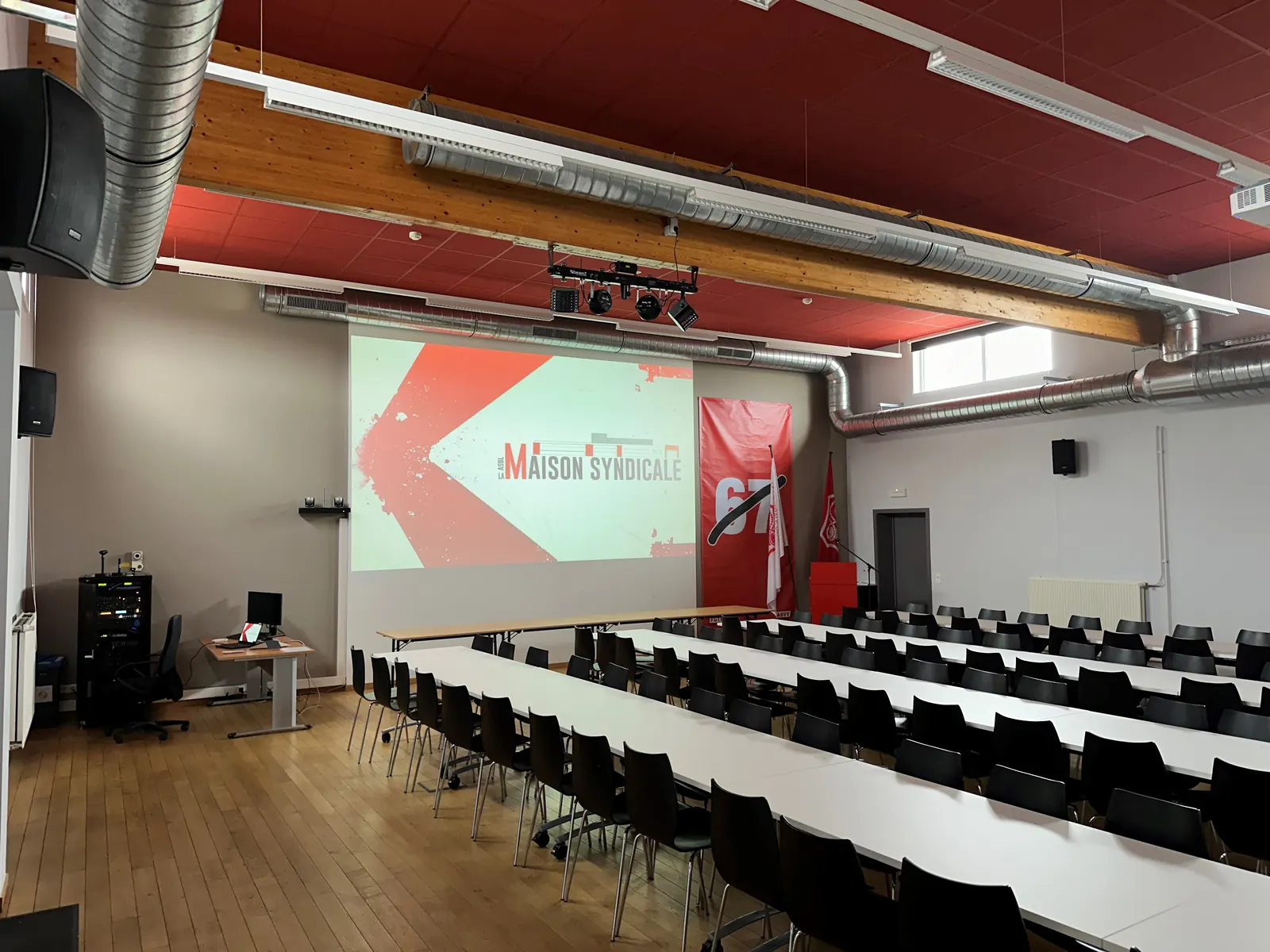 Tables disposées en rangées avec chaises, écran de projection affichant le logo de La Maison Syndicale.