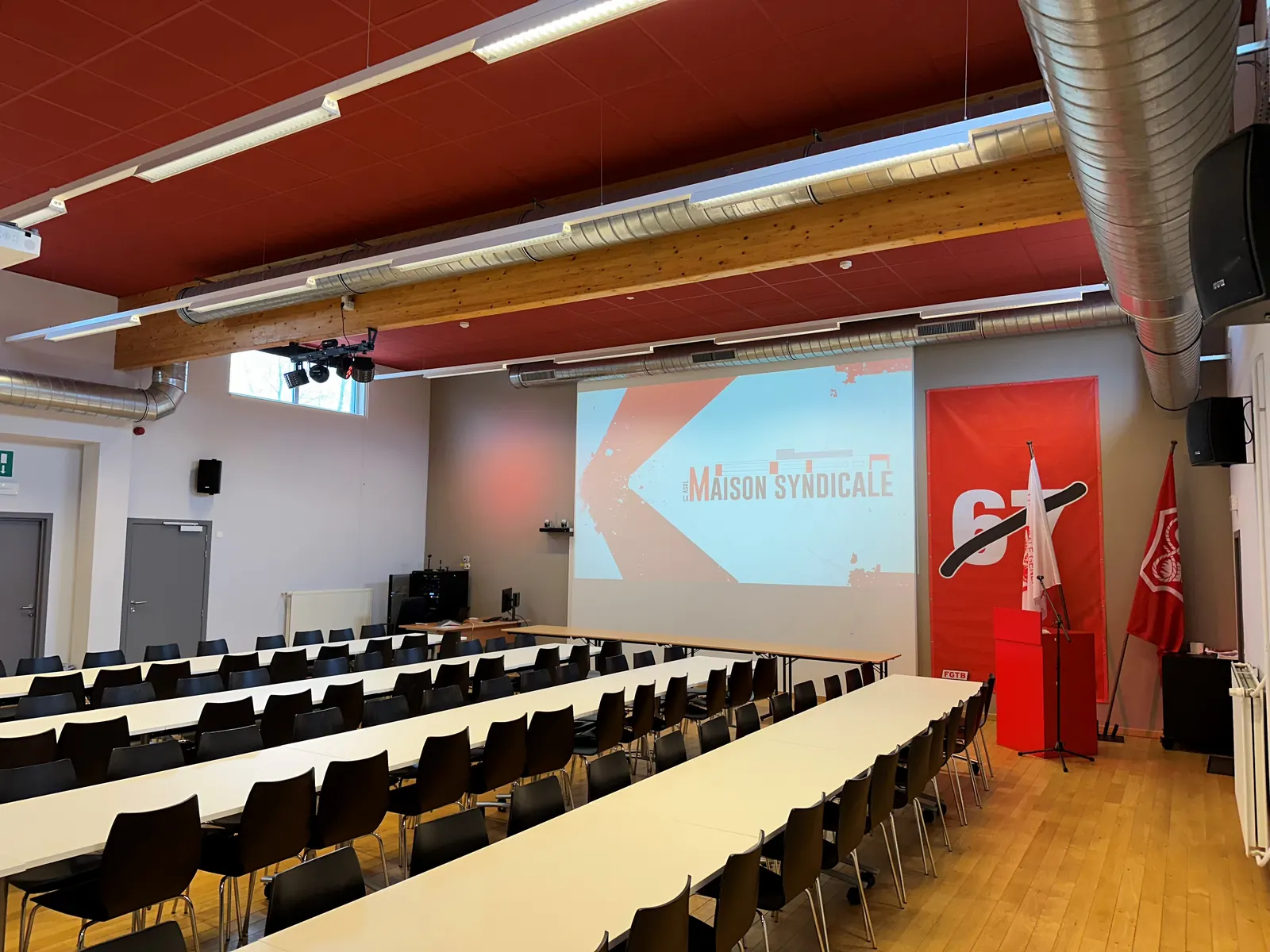 Tables en rangées face à l'écran de projection, drapeaux et bannière rouge à côté de la scène.