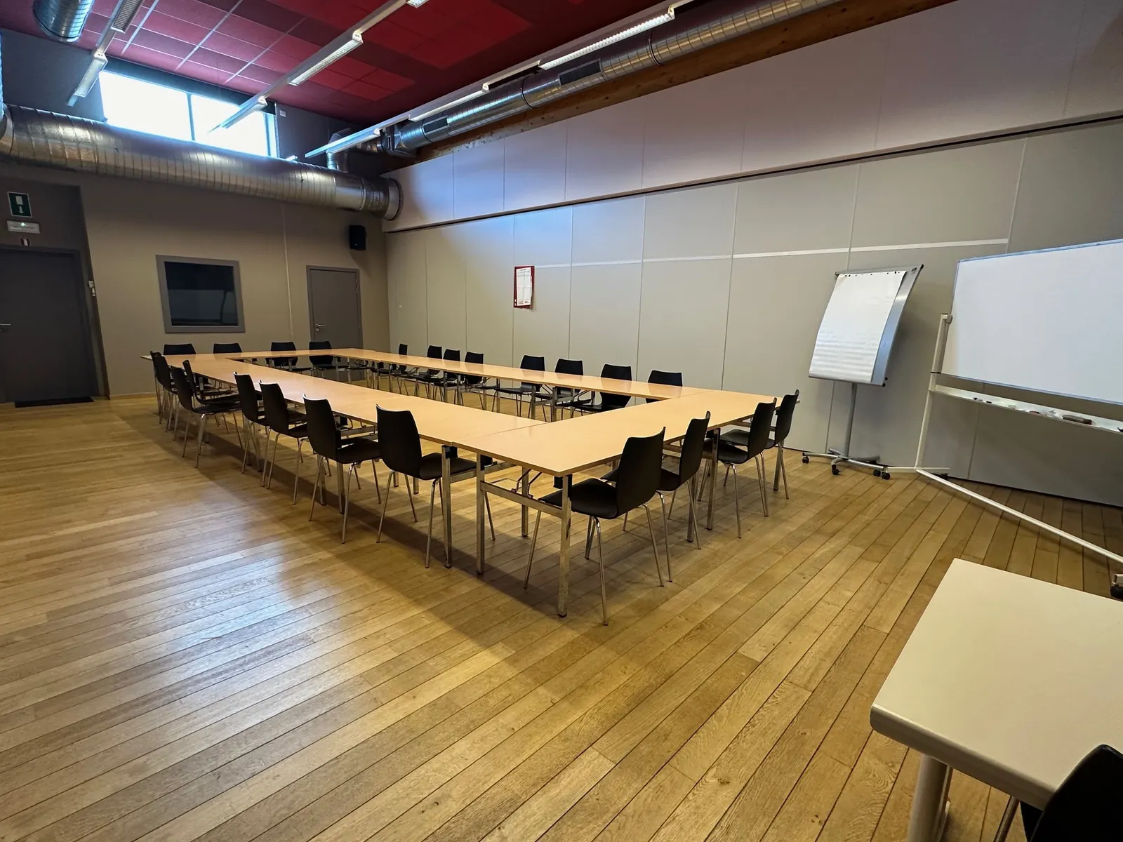 Petite salle : tables disposées en U avec chaises noires, paperboard et tableau blanc, parquet clair.